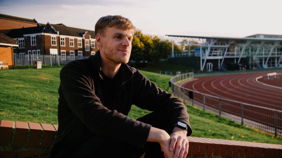 Joe Fuggle smiling into the distance, with the Rutland Building and the University's running track in the background.
